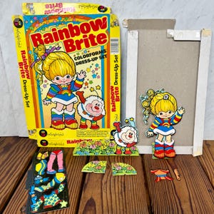 以下が含まれることがあります： ヴィンテージのレインボーブライトカラーフォームズドレスアップセット。箱には、レインボーブライトとスプライトの漫画イラストが描かれています。セットには、ドレスアップパーツと段ボールの背景が含まれています。箱は黄色で、「Rainbow Brite」の文字が赤色で書かれています。