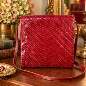 Può includere: Una borsa a tracolla quadrata di colore rosso con un motivo testurizzato in pelle di serpente e linee di cucitura diagonali sulla superficie anteriore. La borsa include una sottile tracolla rossa abbinata e un design a patta strutturato.