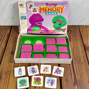 Può includere: Un set scatola vintage di Barney Memory Game. La scatola presenta Barney il dinosauro e un personaggio di dinosauro verde. Il gioco include una scatola, un vassoio con carte rosa e singole carte abbinate con illustrazioni di Barney. La scatola recita "Your First Memory! For Ages 3 to 6."