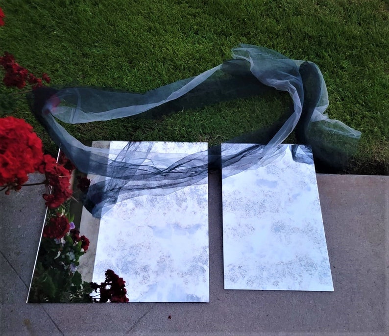 May include: Two rectangular mirrors with a black sheer fabric draped over them. The mirrors are sitting on a concrete surface with green grass in the background.