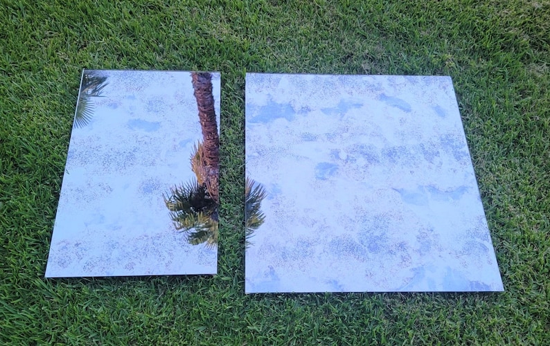 May include: Two rectangular mirrors, one larger than the other, are lying on a green grassy surface. The smaller mirror reflects a palm tree, while the larger mirror reflects a cloudy sky.