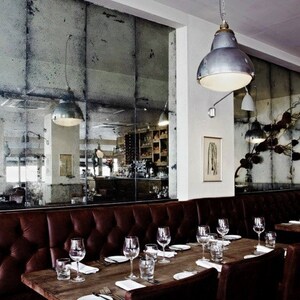 May include: A restaurant interior with a long brown leather banquette, wooden tables, and chairs. The walls are covered in antique mirrored tiles.  The tables are set with glasses, plates, and cutlery.