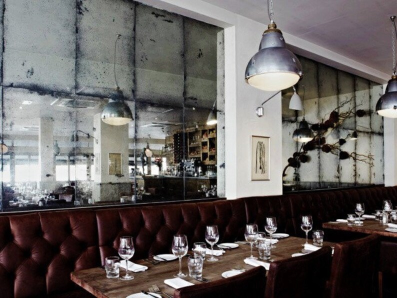May include: A restaurant interior with a long brown leather banquette, wooden tables, and chairs. The walls are covered in antique mirrored tiles.  The tables are set with glasses, plates, and cutlery.