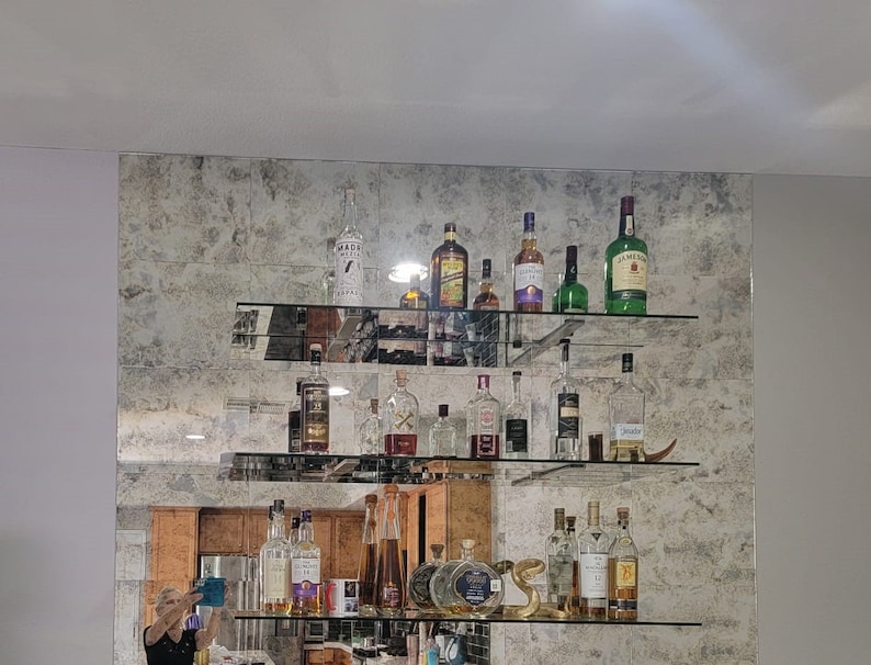 May include: A mirrored wall shelf with multiple glass shelves filled with various bottles of liquor. The bottles are arranged in rows and include brands like Jameson, Maker's Mark, and Patron.