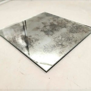 May include: A square piece of antique mirror glass with a distressed, aged look. The mirror has a cloudy, mottled appearance with a silvery sheen.