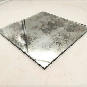May include: A square piece of antique mirror glass with a distressed, aged look. The mirror has a cloudy, uneven surface with a subtle, silvery sheen.