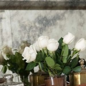 May include: A bouquet of white roses in a clear glass vase on a table with antique-looking silver and gold containers.