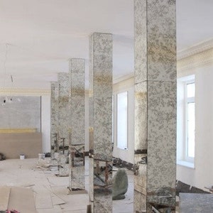 May include: A large room with white walls and a white ceiling. There are several tall, rectangular columns made of mirrored glass. The columns are arranged in a row, and they reflect the room around them. The room is empty, except for some building debris on the floor.