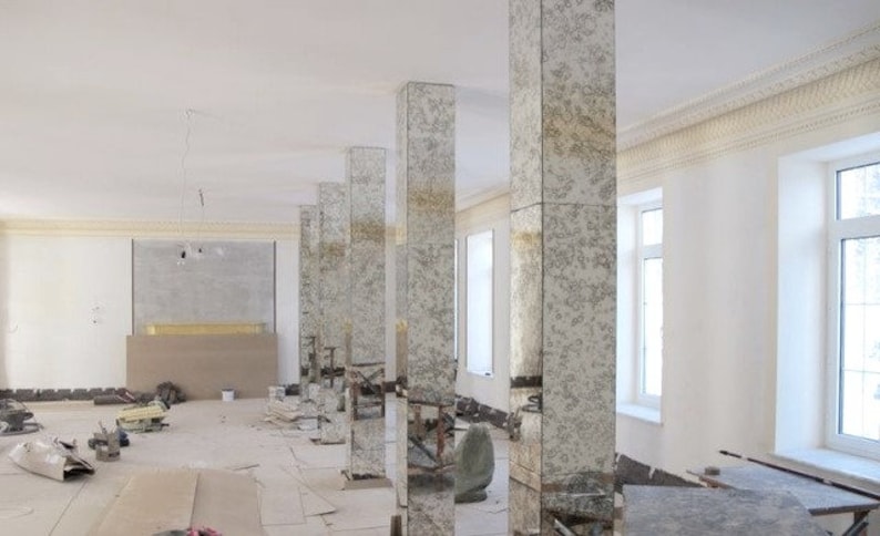 May include: A large room with white walls and a white ceiling. There are several tall, rectangular columns made of mirrored glass. The columns are arranged in a row, and they reflect the room around them. The room is empty, except for some building debris on the floor.
