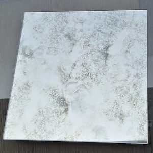 May include: A square, antique mirror with a distressed, silver finish. The mirror has a cloudy, aged appearance with a subtle, mottled pattern.