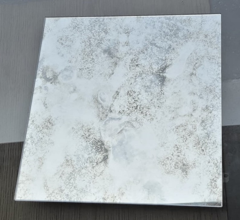 May include: A square, antique mirror with a distressed, silver finish. The mirror has a cloudy, aged appearance with a subtle, mottled pattern.
