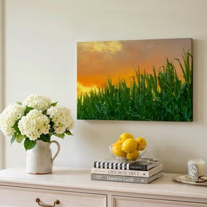 May include: A rectangular canvas wall art print featuring a vibrant sunset sky with orange and yellow hues above a field of green corn stalks. The artwork is displayed on a light-colored wall above a white wooden cabinet with a vase of white flowers.