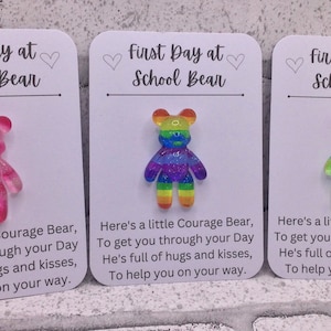 May include: Three cards with the text "First Day at School Bear" and a small, colourful, glittery bear charm on each card. The bears are pink, rainbow, and tie-dye.