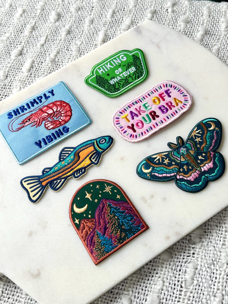Pu&ograve; includere: Collezione di toppe ricamate con vari disegni. Include una toppa blu con un gambero e la scritta "Shrimply Yibing", una toppa verde con "Hiking or Whatever", e una toppa rosa con "Take Off Your Bra". Altre toppe raffigurano un pesce, una scena di montagna e una falena.