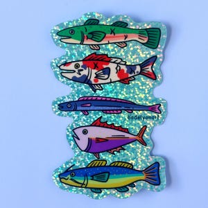 Pode incluir: Cinco adesivos de peixes coloridos em um fundo azul claro. Os peixes são todos de formas e tamanhos diferentes, e todos estão virados para a direita. Os adesivos são feitos de um material brilhante.