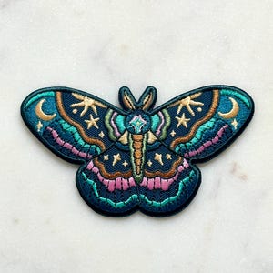 Puede incluir: Parche bordado con un diseño detallado de polilla. La polilla tiene detalles en verde azulado, rosa y dorado, con estrellas y lunas crecientes. El parche está sobre una superficie de mármol blanco.