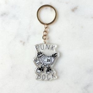 May include: A clear acrylic keychain with a black and white illustration of a punk rock chicken wearing a studded jacket, boots, and a mohawk. The text "Punk Bock" is printed on the keychain.
