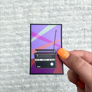 May include: A vintage-style radio with a black and white design, featuring a purple, pink, and orange background with a comic book style pattern. The radio has a white dial with the text "Avent C" and a white knob.