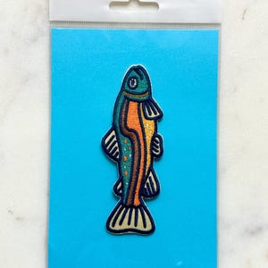 Pu&ograve; includere: Una toppa ricamata a forma di pesce. La toppa presenta una testa verde acqua, un corpo arancione e una coda gialla. Il pesce ha dettagli neri ed &egrave; su una carta blu con un'intestazione bianca.