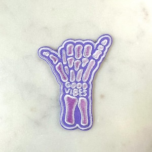 May include: A purple and white embroidered patch in the shape of a hand making the shaka sign. The patch features a skeleton hand design with the words "GOOD VIBES" in the center. The patch is on a white marble surface.