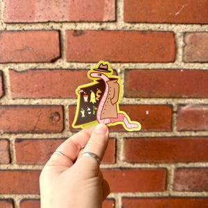 May include: A sticker of a cartoon worm wearing a trench coat and a hat, holding a sign that says "Worms for Sale". The sign has a variety of worms on it, including a red worm, a green worm, a yellow worm, and a brown worm.