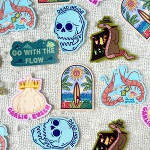 May include: A collection of colorful stickers featuring various designs, including a skull with the text "Dead Inside", a garlic with a crown and the text "Garlic Queen", a worm in a trench coat, and a shrimp with the text "Shrimp Brunch".