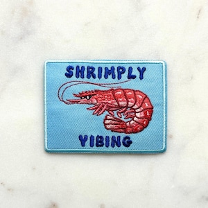 May include: Embroidered patch with a light blue background featuring a red shrimp illustration. The text "SHRIMPLY VIBING" is embroidered in blue above and below the shrimp. The patch has a blue border.