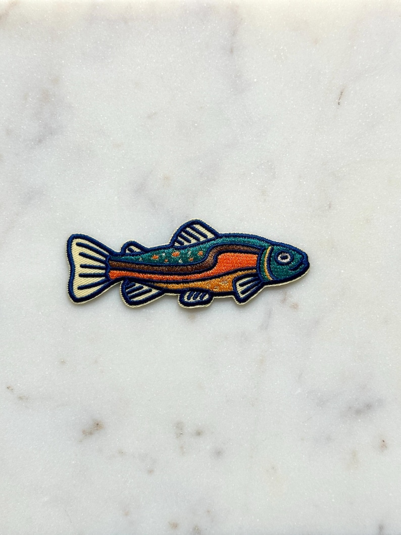 Pu&ograve; includere: Una toppa ricamata colorata a forma di pesce. Il pesce ha una testa blu, un corpo arancione e un dorso verde con piccoli dettagli gialli a forma di stella. La toppa &egrave; bordata di blu scuro e ha pinne color crema.