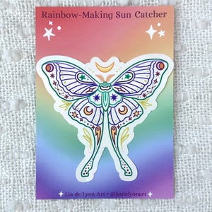 May include: A sticker featuring a white moth with purple and green accents. The moth has a crescent moon on its head and is surrounded by stars. The sticker is on a rainbow background with the text "Rainbow-Making Sun Catcher" and "Lia de Lyon Art @liadel yonart".
