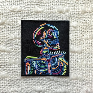 May include: A black patch featuring a neon-coloured skeleton embroidered on it. The skeleton is in profile, with its head tilted to the side. The skeleton is outlined in neon pink, green, blue, and yellow.