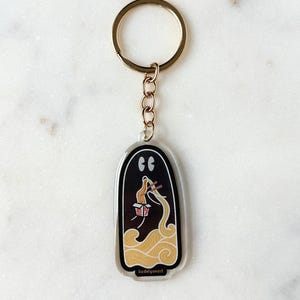 May include: A clear acrylic keychain with a gold-colored ring and chain. The keychain features a black ghost-shaped design with a ramen bowl and noodles. The text "liadelyonart" is printed at the bottom.