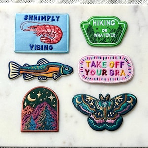 Pu&ograve; includere: Una collezione di toppe ricamate con vari disegni. Una toppa presenta un gambero con il testo "Shrimply Vibing". Un'altra dice "Hiking or Whatever". Altre toppe includono un pesce, una scena di montagna e una farfalla con il testo "Take Off Your Bra."
