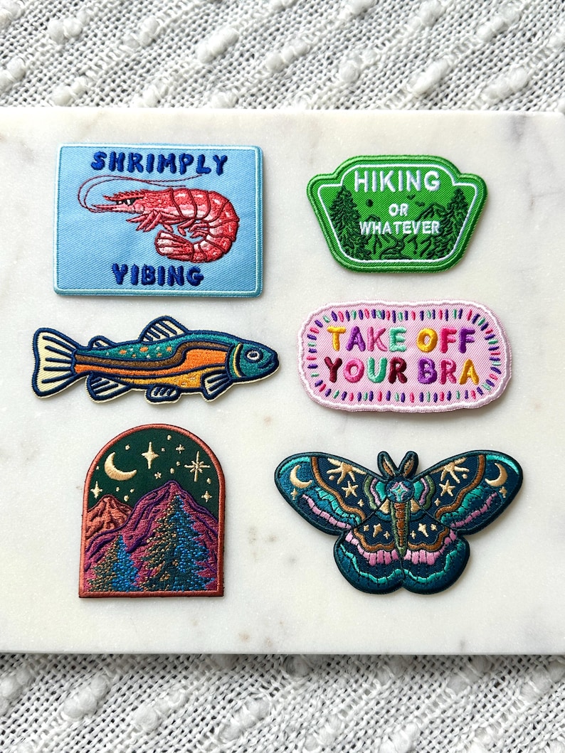 Pu&ograve; includere: Una collezione di toppe ricamate con vari disegni. Una toppa presenta un gambero con il testo "Shrimply Vibing". Un'altra dice "Hiking or Whatever". Altre toppe includono un pesce, una scena di montagna e una farfalla con il testo "Take Off Your Bra."