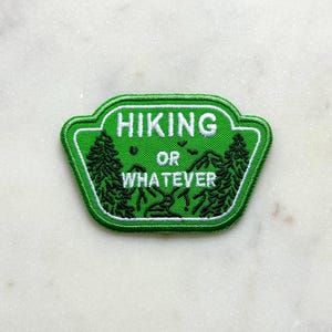 May include: Green embroidered patch with the words "HIKING OR WHATEVER" in white. The patch features a mountain scene with trees and silhouettes of people hiking. The patch has a white border.
