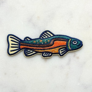 May include: Embroidered patch in the shape of a fish. The fish has a blue head and tail, with orange and brown body markings. The patch is outlined in a cream color and is likely for clothing or accessories.
