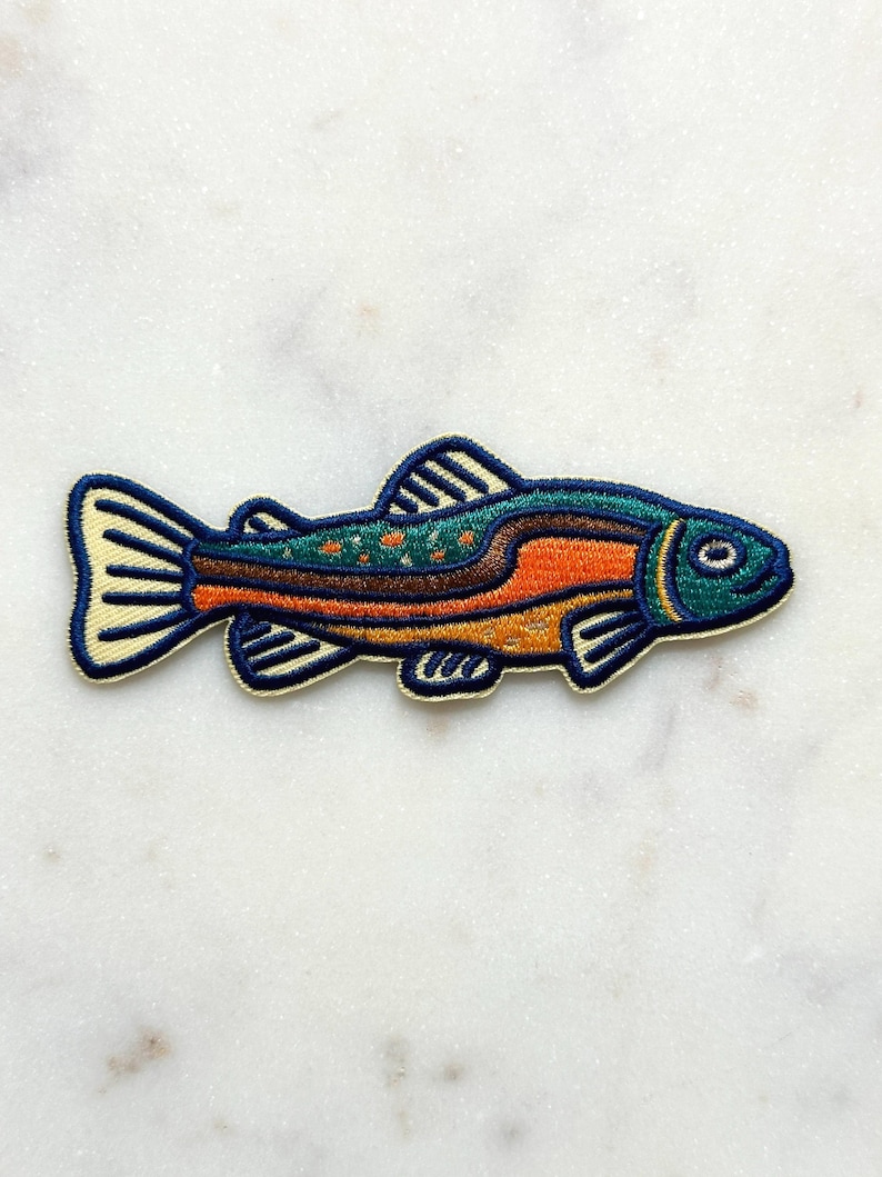 Pu&ograve; includere: Toppa ricamata a forma di pesce. Il pesce ha testa e coda blu, con segni arancioni e marroni sul corpo. La toppa &egrave; bordata di color crema ed &egrave; probabilmente per abbigliamento o accessori.