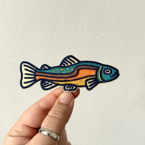 Pu&ograve; includere: Toppa ricamata con un design di pesce stilizzato. Il pesce ha una testa turchese, un corpo arancione e giallo e un contorno blu navy. La toppa &egrave; tenuta da una mano su uno sfondo neutro.