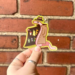 May include: A pink worm wearing a brown trench coat and a hat, holding a sign that says "WORM" and has images of various food scraps on it. The background is a brick wall.