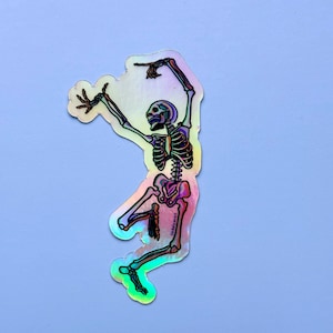 Puede incluir: Una pegatina holográfica de un esqueleto bailando con los brazos en alto. La pegatina es iridiscente y tiene un brillo arcoíris. El esqueleto está delineado en negro y tiene una caja torácica y un cráneo detallados.