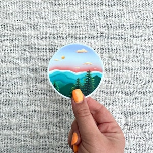 Puede incluir: Una pegatina redonda con una ilustración colorida de una cordillera con un cielo de puesta de sol y nubes. Las montañas son verdes, azules y moradas, y el cielo es rosa, naranja y amarillo.