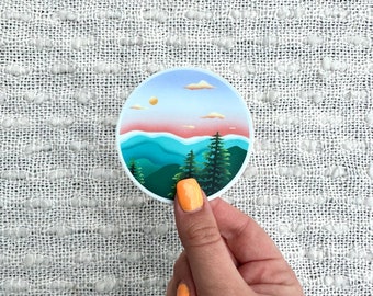 May include: A round sticker with a colorful illustration of a mountain range with a sunset sky and clouds. The mountains are green, blue, and purple, and the sky is pink, orange, and yellow.