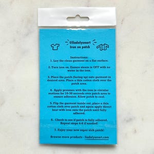 Pu&ograve; includere: Un pacchetto di toppa termoadesiva turchese con istruzioni. Il pacchetto include il testo "@liadelyonart Iron on patch" e le istruzioni per l'applicazione. Lo sfondo &egrave; una superficie di marmo bianco.