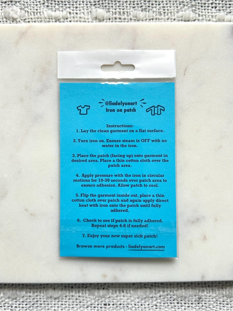 Pu&ograve; includere: Un pacchetto di toppa termoadesiva turchese con istruzioni. Il pacchetto include il testo "@liadelyonart Iron on patch" e le istruzioni per l'applicazione. Lo sfondo &egrave; una superficie di marmo bianco.