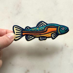 Pu&ograve; includere: Toppa ricamata a forma di pesce con un design colorato. La toppa ha una testa e una coda blu, una sezione centrale arancione e un dorso turchese con pois arancioni. Il pesce ha un contorno nero e dettagli gialli.