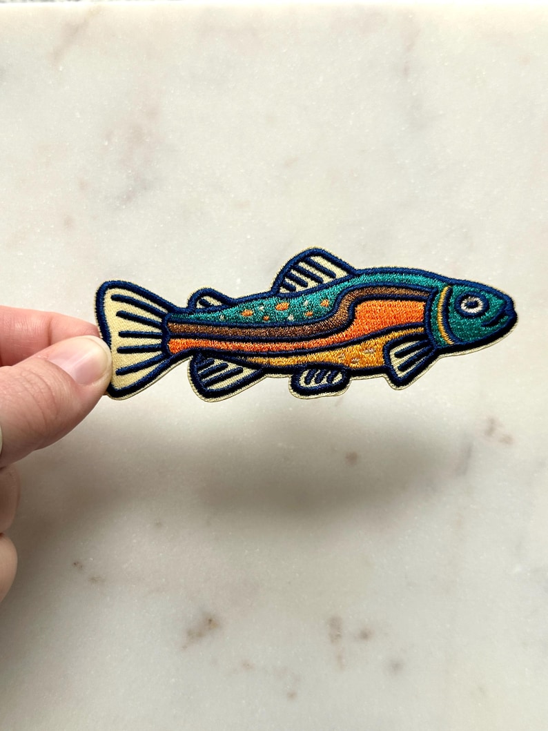 Pu&ograve; includere: Toppa ricamata a forma di pesce con un design colorato. La toppa ha una testa e una coda blu, una sezione centrale arancione e un dorso turchese con pois arancioni. Il pesce ha un contorno nero e dettagli gialli.