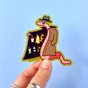 May include: A cartoon worm wearing a brown trench coat and a fedora holds a sign that says "I WAS", with a variety of fruit peels and cores on the sign. The worm is pink and yellow and has a black eye. The background is light blue.