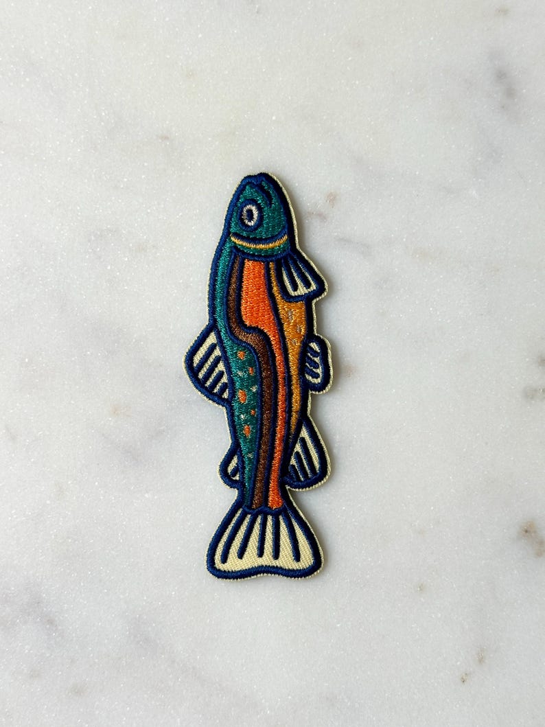 Pu&ograve; includere: Toppa ricamata a forma di pesce. Il pesce ha una testa turchese, un corpo arancione e dettagli blu e gialli. Adatto per abbigliamento o accessori.