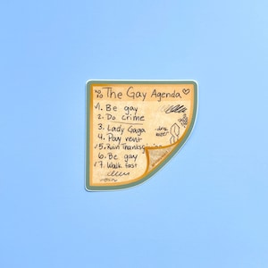 Op de afbeelding: Een sticker op een lichtblauwe achtergrond met een handgeschreven lijst getiteld "The Gay Agenda". De lijst bevat items als "Be gay", "Do crime" en "Lady Gaga". De sticker heeft een bruingeel achtergrond met een gebogen rand.