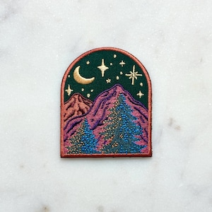 May include: Embroidered patch depicting a night scene with mountains, stars, and a crescent moon. The patch has a dark green background with a red border. The mountains are purple and the trees are blue and gold.