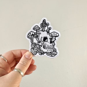 May include: Black and white embroidered patch of a skull with mushrooms growing out of its head.